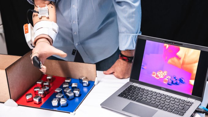 A man's prosthetic hand hovers over metal cubes that he sorted into a red area for hot and blue area for cold. A sensor on the index finger of the prosthetic hand is connected to a box higher up on his arm where the nerve impulses to sense temperature originate. A thermal image on a laptop show that the cubes were sorted correctly.