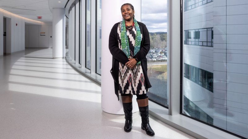 A photograph of Oluwatoyin Asojo who's faintly smiling while standing in an empty white hallway by large panels of windows. She is wearing a dress with black, white, brown and red geometric patterns, black coat, black and brown knee-high boots, green scarf with patterns, and brown and orange necklace.