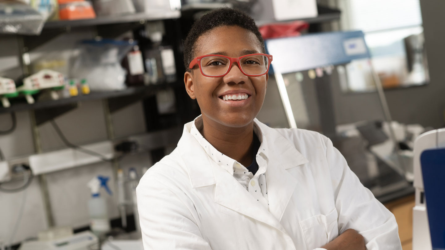 Dionna Williams is smiling at the camera. They have short, dark hair and are wearing orange-rimmed eyeglasses and white lab coat. They are standing in a lab with their arms crossed.