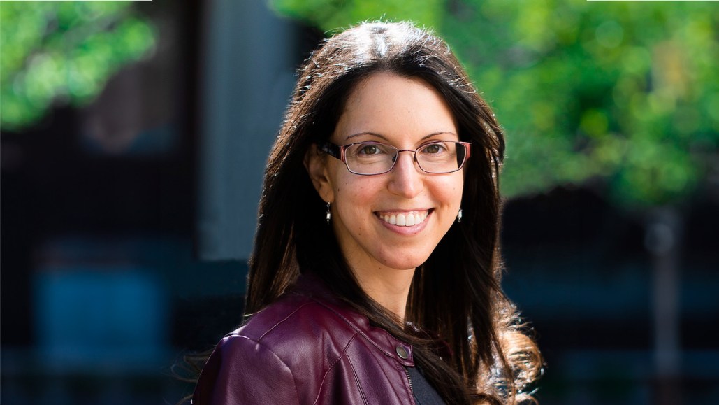 Nicole Yunger Halpern is looking at the camera while smiling widely. She has long brown hair and wears glasses.