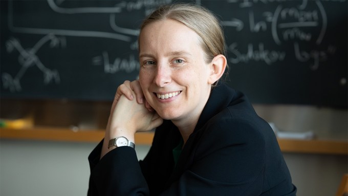 Tracy Slatyer is staring at the camera, smiling. There is a black board with equations written on in the background. Slatyer is wearing a black jacket and silver watch. Her light brown (or dark blonde) hair is pulled back in a ponytail.